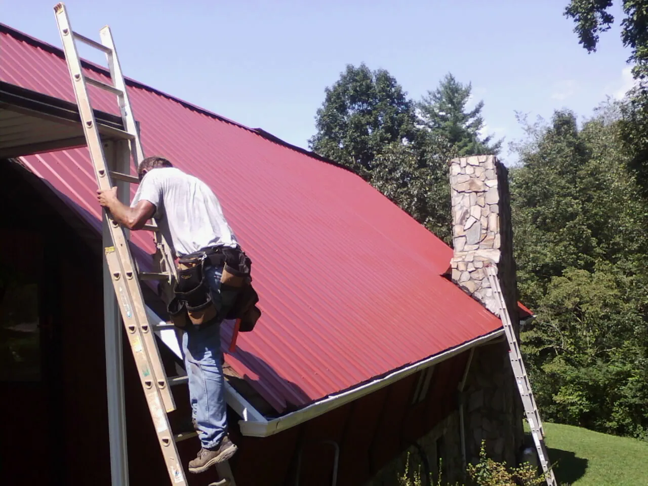Roofer on a ladder beside a red metal roof and stone chimney — Soffit & Fascia Repair by Roofing Brothers in Salem